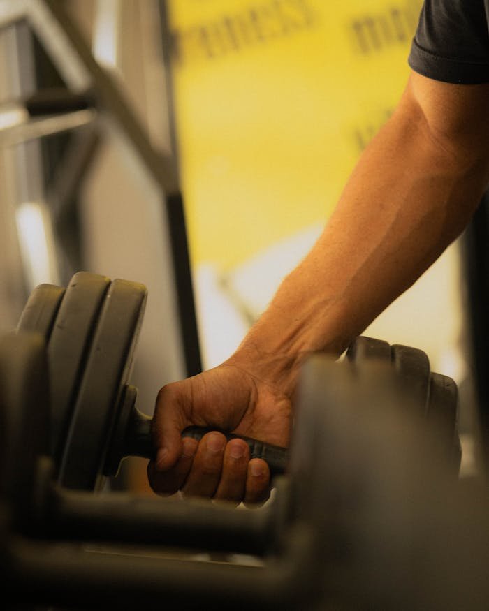 gallery-2 Close-up of a muscular arm gripping a dumbbell, emphasizing strength and power inside a gym.