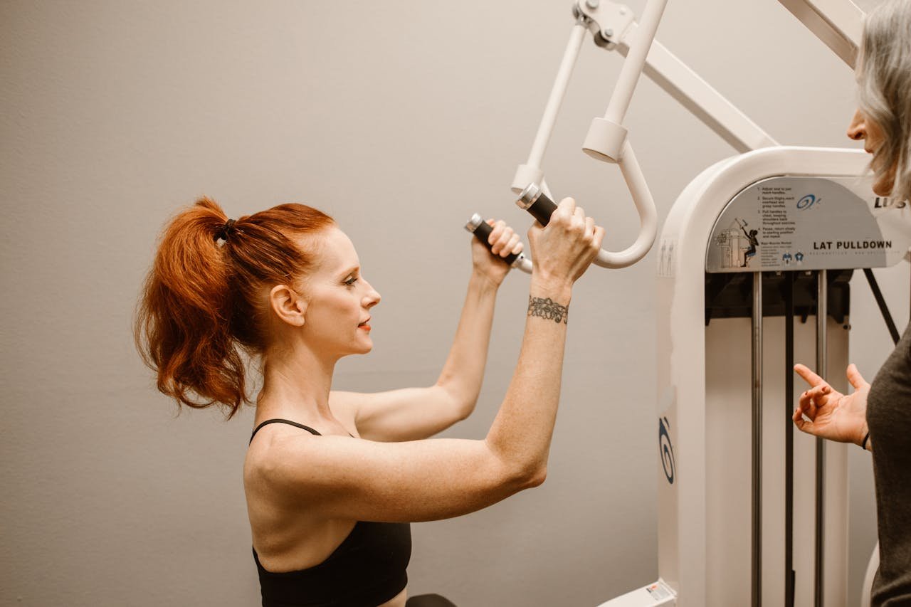 gallery-1 Woman working out on lat pulldown machine while being guided by a trainer.