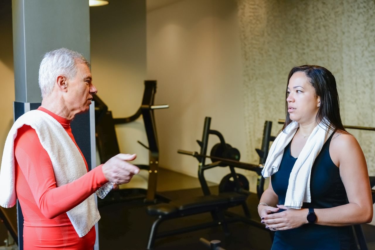 hero-img-01 An elderly man and woman discuss fitness routines in a gym setting.